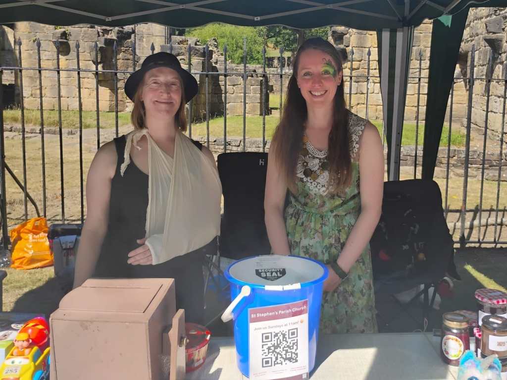 Two women smiling at the camera, standing in Kirkstall abbey, behind a donations bucket that says St Stephen's Church.